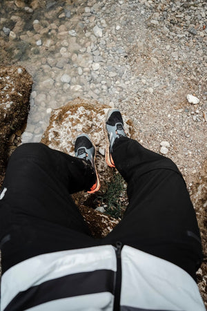 Point-of-view shot looking down at a hiker's muddy gray trail shoes with orange soles, resting on a rock beside a pebbly stream.