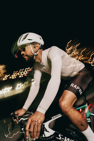 Man riding a bike at night, wearing a full cycling kit with black Rapha shorts, a white thermo shirt, cycling glasses and a white helmet. Picture by Venti Views in Unsplash.